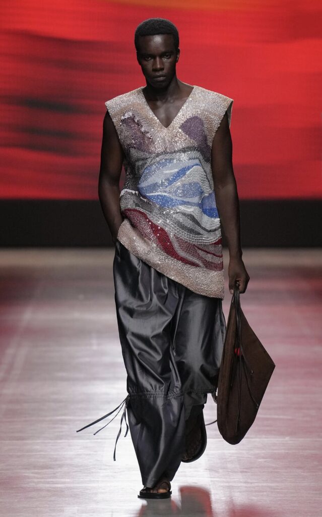 A male model walking down the runway wearing a delicate sequinned and patterned vest with black leather pants.